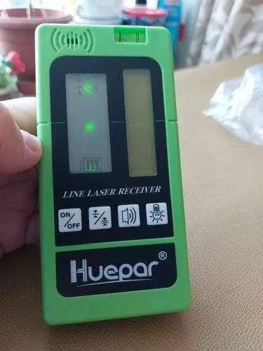Green Huepar line laser receiver with display lights and control buttons, shown handheld indoors.